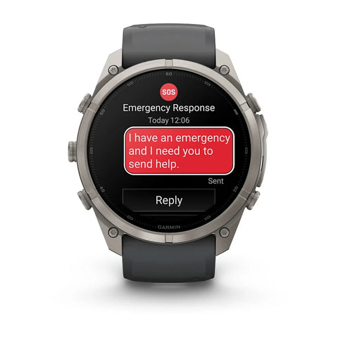 A Garmin smartwatch with a dark gray strap and a silver-toned metal case, shown on a clean white background. The watch screen displays an emergency message with a red SOS icon and a red text bubble reading 'I have an emergency and I need you to send help.