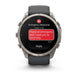 A Garmin smartwatch with a dark gray strap and a silver-toned metal case, shown on a clean white background. The watch screen displays an emergency message with a red SOS icon and a red text bubble reading 'I have an emergency and I need you to send help.