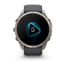 A Garmin smartwatch with a dark gray silicone strap and a silver-toned metallic case, shown against a plain white background. The watch face is black with a glowing blue geometric icon in the center.