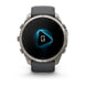 A Garmin smartwatch with a dark gray silicone strap and a silver-toned metallic case, shown against a plain white background. The watch face is black with a glowing blue geometric icon in the center.