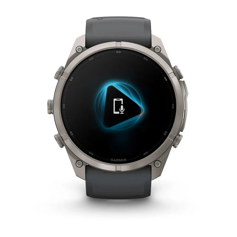 A Garmin smartwatch with a dark gray silicone strap and a silver-toned metallic case, shown against a plain white background. The watch face is black with a glowing blue geometric icon in the center.