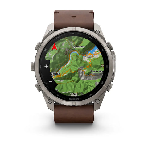 A Garmin smartwatch with a round silver-tone metal case and a brown leather strap, shown on a clean white background. The watch face displays a green topographic map.