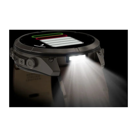A rugged smartwatch with a circular display and angular side buttons, shown in profile with its front light turned on and casting a bright beam of light downward. The watch has a dark gray and black body with a light gray strap, set against a black backgr