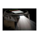 A rugged smartwatch with a circular display and angular side buttons, shown in profile with its front light turned on and casting a bright beam of light downward. The watch has a dark gray and black body with a light gray strap, set against a black backgr