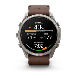 A Garmin smartwatch with a silver-toned metal case and a brown leather strap, shown on a clean white background. The watch face is active with various fitness and heart rate metrics displayed.
