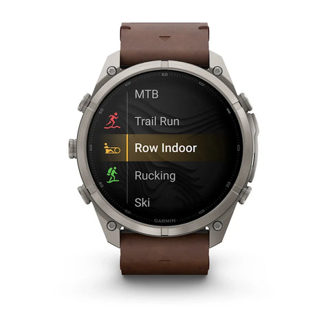 A Garmin smartwatch with a silver-toned metal case and a brown leather strap, shown on a clean white background. The watch face displays a fitness menu with options like MTB, Trail Run, Row Indoor, Rucking, and Ski.