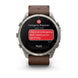 A Garmin smartwatch with a silver-toned metal case and a brown leather strap, shown on a clean white background. The watch screen displays an emergency message interface with a red SOS badge, a red message bubble, and a black background.