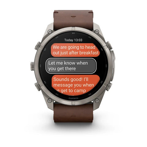 A Garmin smartwatch with a silver-toned metal case and a brown leather strap, shown on a clean white background. The watch screen displays a messaging app interface with orange and gray text bubbles.