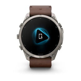 A Garmin smartwatch with a round silver-tone metal case and a brown leather strap, shown against a plain white background. The watch face is black with a glowing blue geometric icon in the center.