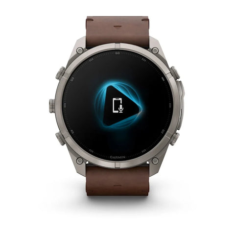A Garmin smartwatch with a round silver-tone metal case and a brown leather strap, shown against a plain white background. The watch face is black with a glowing blue geometric icon in the center.