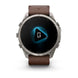 A Garmin smartwatch with a round silver-tone metal case and a brown leather strap, shown against a plain white background. The watch face is black with a glowing blue geometric icon in the center.