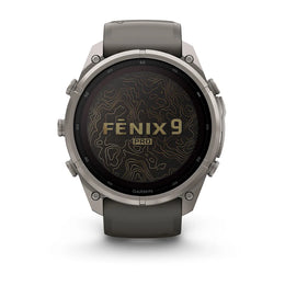 A Garmin Fenix 9 Pro smartwatch with a round silver-toned metal case, dark gray strap, and a black display showing a topographic map pattern with gold lettering.