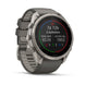 A Garmin smartwatch with a dark gray strap and a silver-toned metal case, shown at a three-quarter angle on a clean white background. The watch face displays fitness metrics.