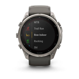 A Garmin smartwatch with a round silver-tone metal case and a dark gray silicone strap, shown against a plain white background. The watch face is on and displaying a list of activities including MTB, Trail Run, Row Indoor, Rucking, and Ski.