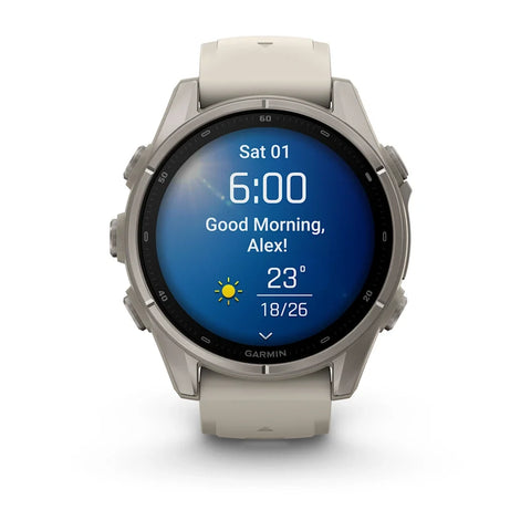A Garmin smartwatch with a round silver stainless steel case and a light beige silicone strap, shown on a clean white background. The watch face is illuminated with a blue digital weather display.