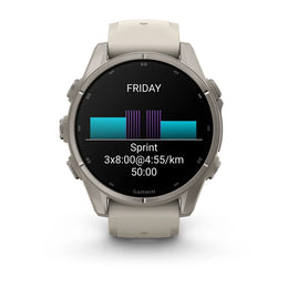 A Garmin smartwatch with a round silver-toned metal case and a light beige silicone strap, shown on a clean white background. The watch face is active with a black display showing a fitness graphic and the word 'FRIDAY'.