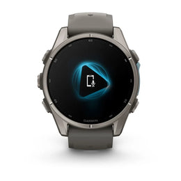 A Garmin smartwatch with a round metallic gray case and a dark gray strap, shown against a plain white background. The watch face displays a black screen with a glowing blue geometric icon and a white microphone symbol.