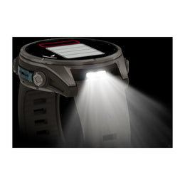 A rugged smartwatch with a dark strap and metallic case, shown with its front light turned on and emitting a bright beam of light downward. The watch face displays a battery graphic, and the side of the watch has a blue accent.