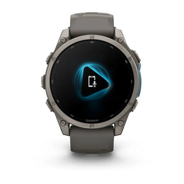A Garmin smartwatch with a dark gray silicone strap and a metallic gray round case, shown against a plain white background. The watch face is black with a glowing blue geometric icon in the center.