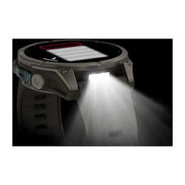 A rugged smartwatch with a circular display and side button, shown in a dark setting with its front light turned on, casting a bright beam of light forward. The watch face displays a battery icon.