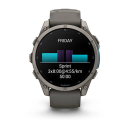 A Garmin smartwatch with a dark gray silicone strap and a metallic dark gray bezel, shown on a clean white background. The watch face displays a fitness screen with a cycling or running split graphic and the word 'FRIDAY'.