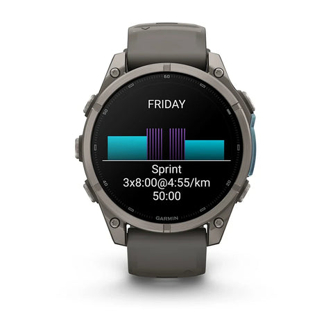 A Garmin smartwatch with a dark gray silicone strap and a metallic dark gray bezel, shown on a clean white background. The watch face displays a fitness screen with a cycling or running split graphic and the word 'FRIDAY'.