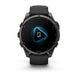 A Garmin smartwatch with a round dark gray metallic case and a black strap, shown from the front on a white background. The watch face displays a black screen with a glowing blue geometric icon in the center.