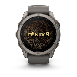 A Garmin Fenix 9 smartwatch with a dark gray titanium case and a dark gray strap, featuring a black screen with gold topographic lines and the text 'FENIX 9' in gold. The watch is centered on a clean white background.