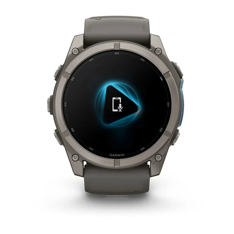 A rugged smartwatch with a dark gray metallic case and matching dark gray strap, shown from the front on a clean white background. The watch face displays a black screen with a glowing blue geometric icon and the Garmin logo at the bottom.