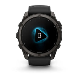 A black Garmin smartwatch with a round dark gray case and a black strap, shown from the front on a white background. The watch screen displays a blue and white audio interface.