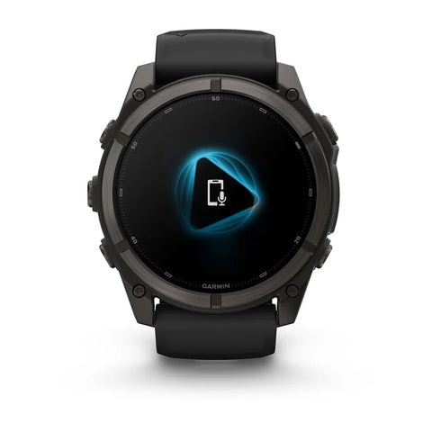 A black Garmin smartwatch with a round dark gray case and a black strap, shown from the front on a white background. The watch screen displays a blue and white audio interface.