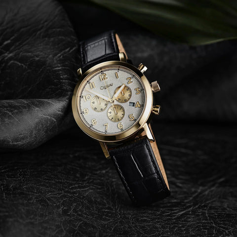 A luxury chronograph watch with a gold-tone case, white dial, and black leather strap, resting on a textured black surface.