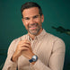 A smiling man with short brown hair and a light beard wears a beige and white vertically striped button-down shirt and a dark blue watch with a silver-toned case, posed against a dark green background.