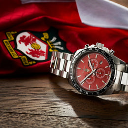 A stainless steel chronograph watch with a red dial and black bezel, resting on a wooden surface in front of a red and white flag with a crest.