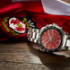 A stainless steel chronograph watch with a red dial and black bezel, resting on a wooden surface in front of a red and white flag with a crest.