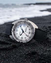 A DOWA Proxima watch with a silver stainless steel case, white dial, and black textured strap, resting on a black sand beach with the ocean waves blurred in the background.