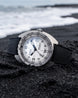 A DOWA Proxima watch with a silver stainless steel case, white dial, and black textured strap, resting on a black sand beach with the ocean waves blurred in the background.