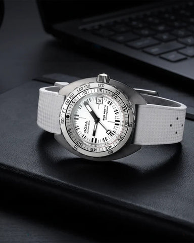 A wristwatch with a white textured strap, a silver-tone case, and a white dial with black markings, resting on a black surface with a blurred laptop keyboard in the background.