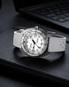 A wristwatch with a white textured strap, a silver-tone case, and a white dial with black markings, resting on a black surface with a blurred laptop keyboard in the background.