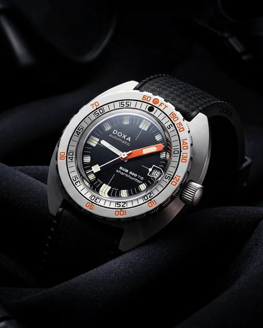 A DOXA automatic wristwatch with a black dial, orange accents on the bezel and hands, and a black textured strap, resting on dark draped fabric.