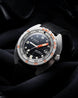 A DOXA automatic wristwatch with a black dial, orange accents on the bezel and hands, and a black textured strap, resting on dark draped fabric.