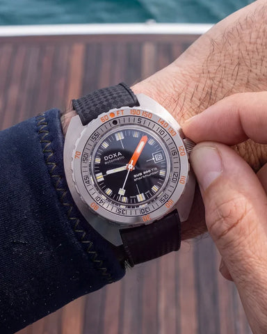 A close-up of a person's wrist wearing a DOXA diver watch with a black dial, black strap, and a silver-toned bezel featuring orange markings, with a hand adjusting the crown.