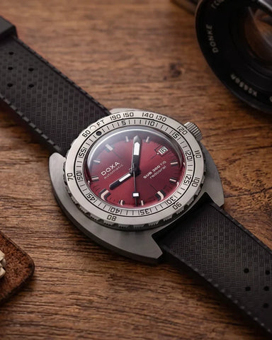 A close-up of a Doxa wristwatch with a dark red dial and a textured black strap, resting on a rustic wooden surface with a camera lens partially visible in the background.
