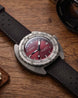 A close-up of a Doxa wristwatch with a dark red dial and a textured black strap, resting on a rustic wooden surface with a camera lens partially visible in the background.