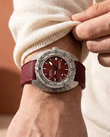 A close-up of a wrist wearing a dialux wrist watch with a textured burgundy leather strap and a matching burgundy dial, paired with a cream knit sweater.