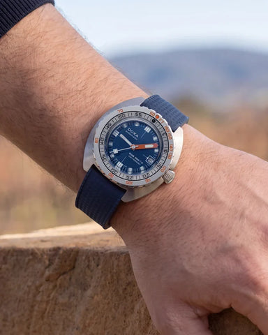 A wristwatch with a blue dial, silver-toned case, and dark blue fabric strap, worn on a person's wrist against a blurred outdoor background.