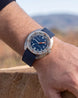 A wristwatch with a blue dial, silver-toned case, and dark blue fabric strap, worn on a person's wrist against a blurred outdoor background.