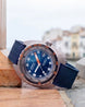 A Doka dive watch with a blue dial, blue strap, and silver-toned case, resting on a stone ledge with a blurred coastal town background.