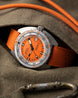 A close-up of a Kova dive watch with a bright orange dial and matching orange strap, resting on a textured olive-brown fabric surface with an orange cord and metal clasp visible.