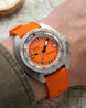 A close-up of a DOXA 200m Automatic watch with a bright orange dial and matching orange textured strap, worn on a wrist over an olive green sleeve.